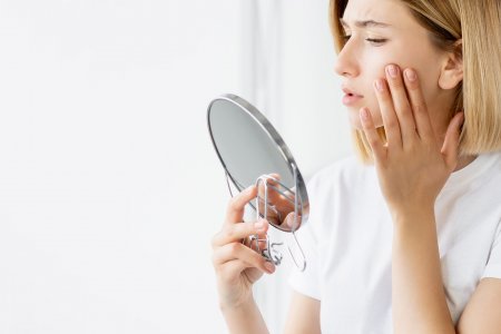 Moteris veidrodyje apžiūri savo spuoguotą odą ir džiaugiasi pagerėjusia būkle laikydamasi švarios odos procedūrų.